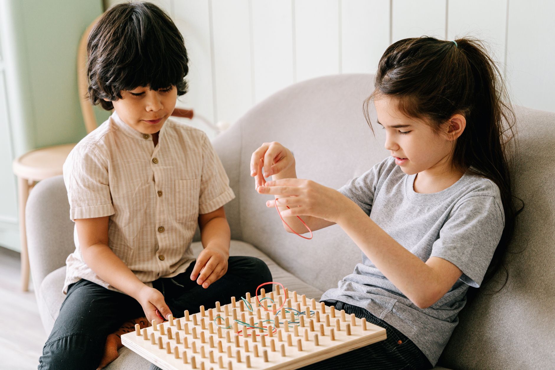 Deux enfants assis sur un sofa jouent avec des élastiques sur une planche en bois munie de picots.