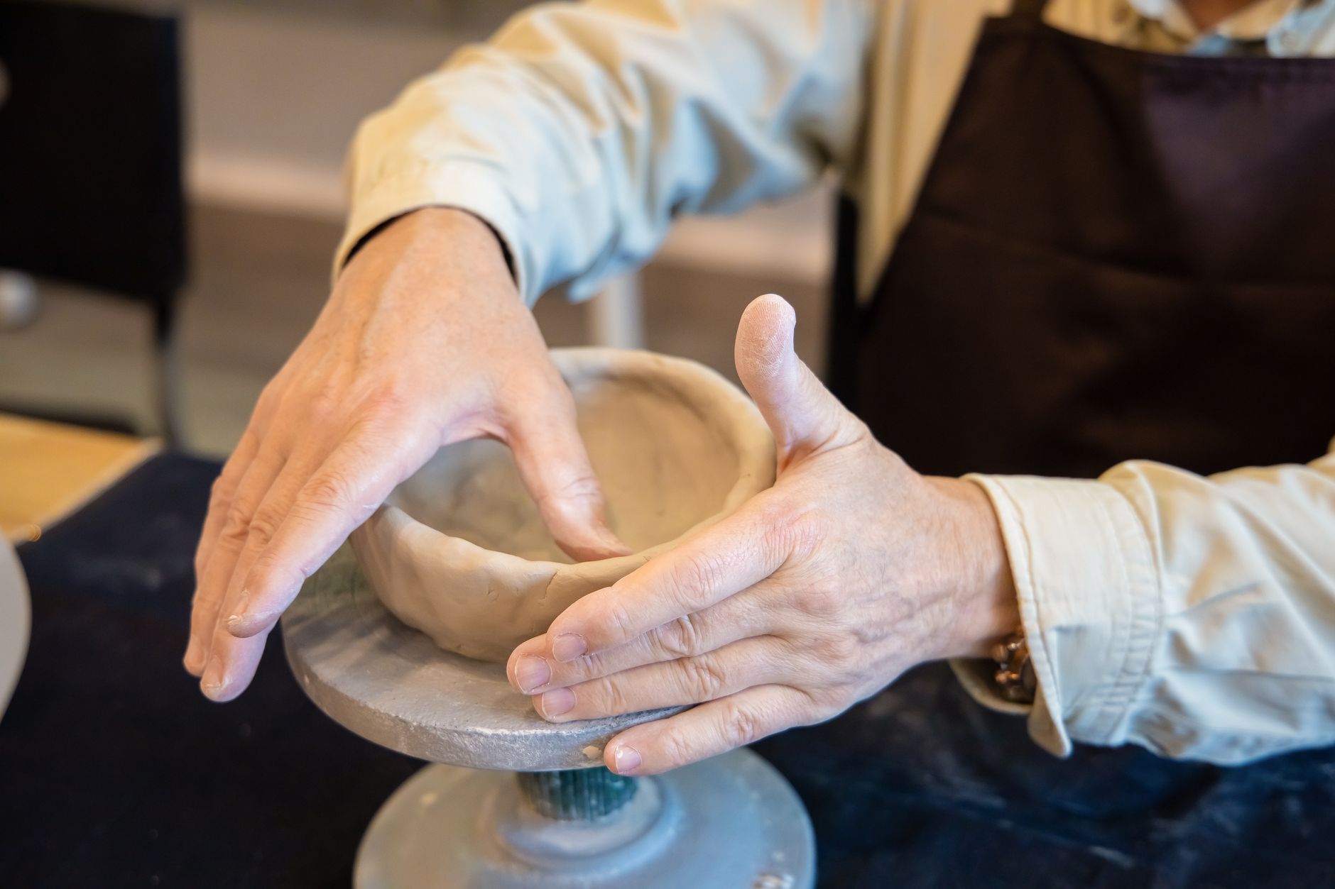 Deux mains façonnent un bol d’argile posé sur un support de poterie, devant une personne en chemise claire et tablier foncé.