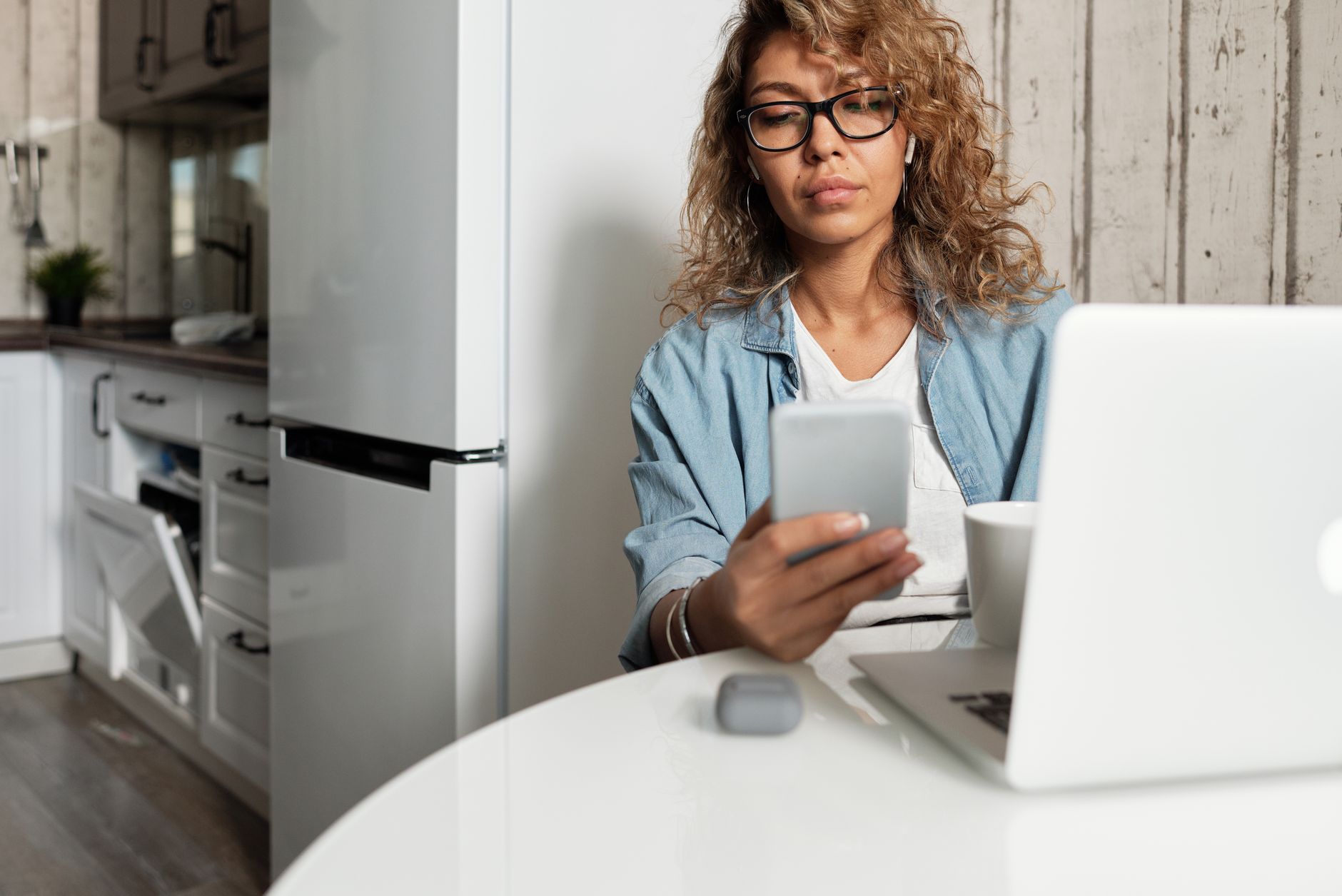Femme à lunettes consultant son smartphone à une table de cuisine, avec un ordinateur portable et une tasse devant elle.