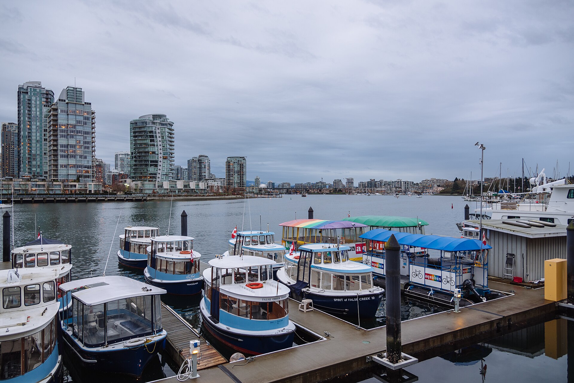 Petits bateaux-taxis bleus et blancs amarrés à un ponton de Granville Island, face à des tours résidentielles