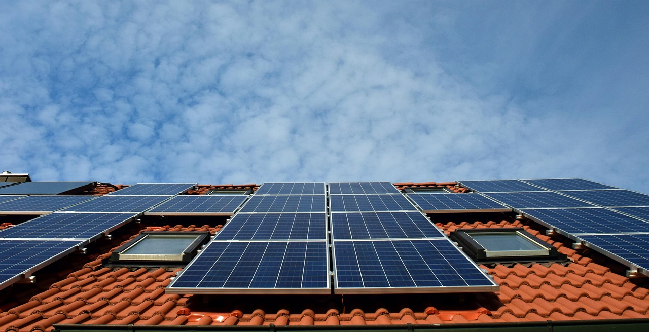 Rangées de panneaux photovoltaïques et fenêtres de toit sur une toiture en tuiles rouges sous un ciel nuageux.