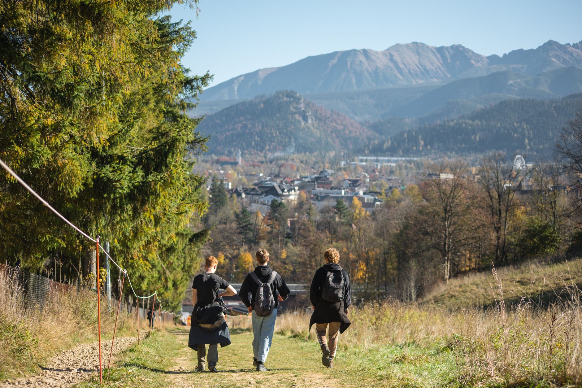 Trois personnes de dos, sac au dos, marchent sur un chemin herbeux vers un village de vallée entouré de montagnes en automne.