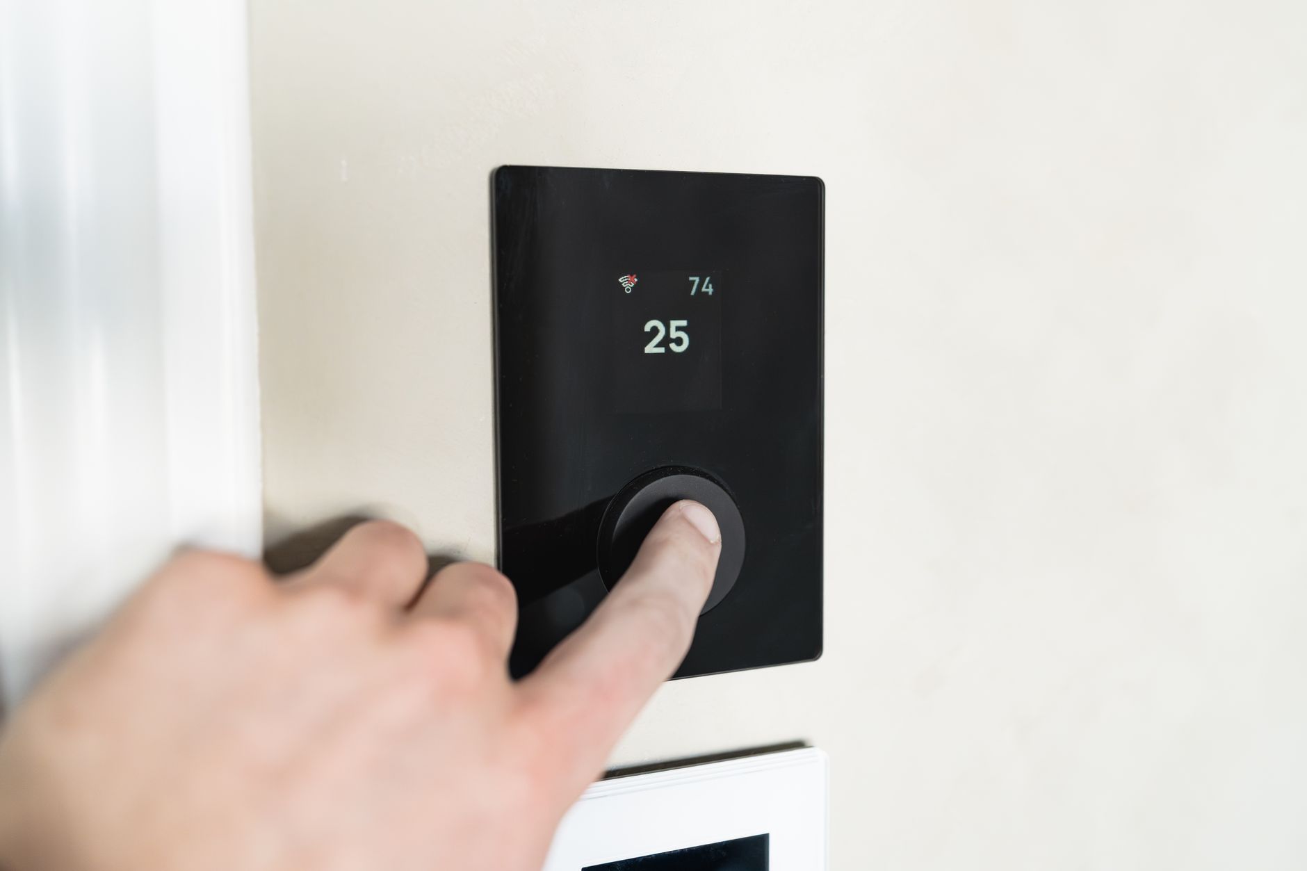 Une main appuie sur le bouton d’un thermostat mural noir dont l’écran affiche 25, installé sur un mur beige clair.